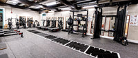 A spacious indoor gym with treadmills, weight machines, punching bags, and black rubber training mats laid out on a gray floor. The space has an industrial ceiling with exposed beams and fluorescent lighting, and white walls with framed equipment along one side.