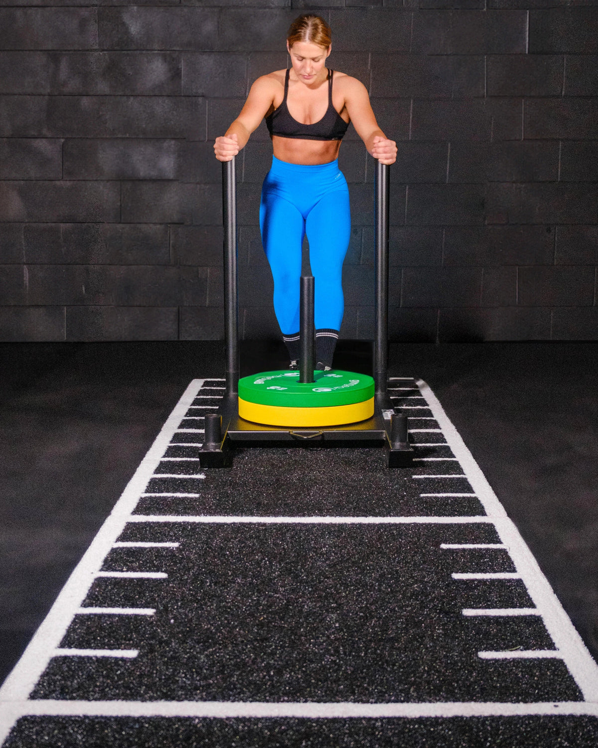 A woman in  bright blue leggings stands behind a black sled exercise machine with a green and yellow weight plate, on a black gym floor with white track markings against a dark brick wall.