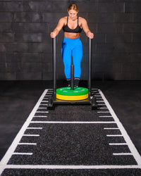 A woman in  bright blue leggings stands behind a black sled exercise machine with a green and yellow weight plate, on a black gym floor with white track markings against a dark brick wall.