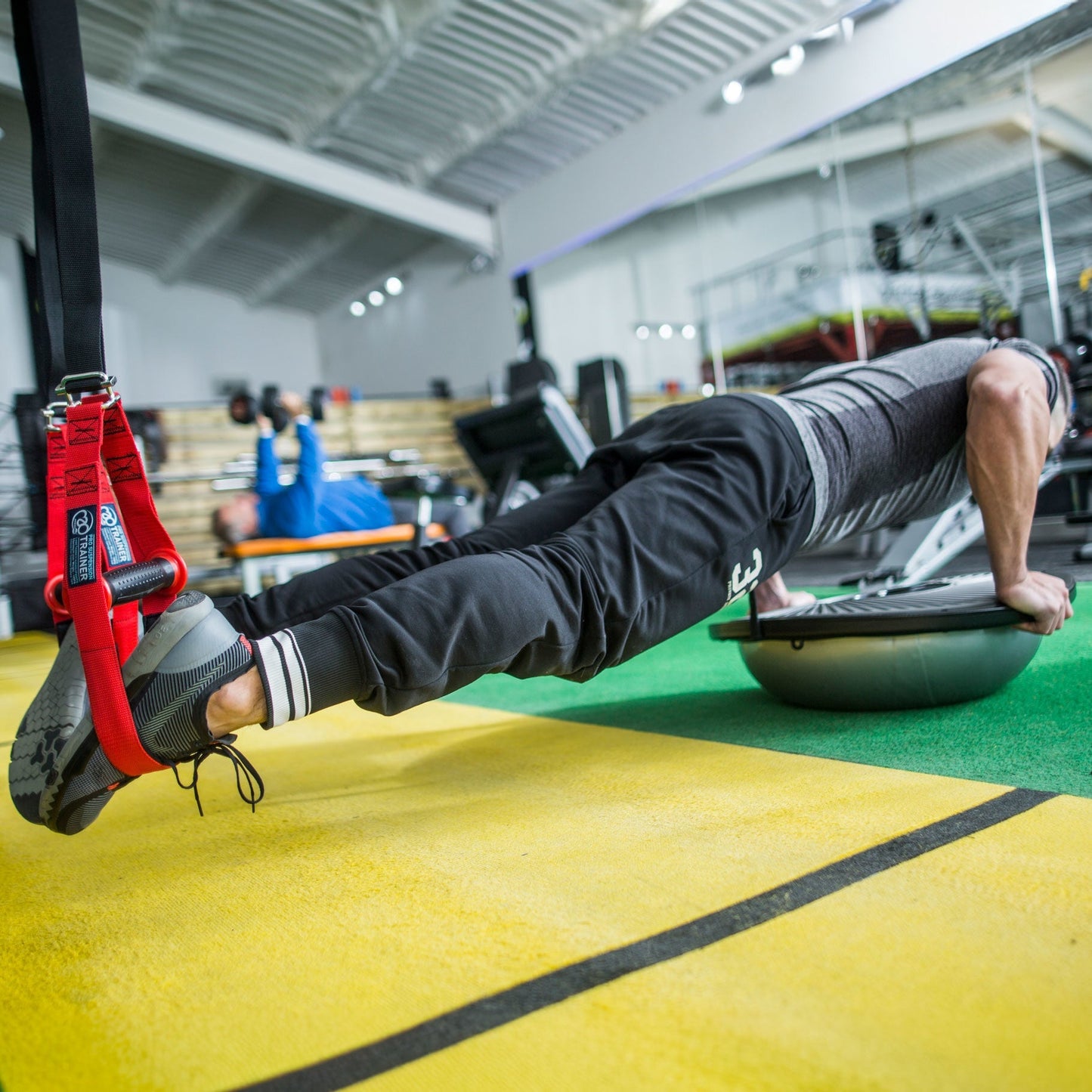 A person in a gym performing a push-up on a BOSU ball, with red suspension straps hanging at the left edge.