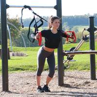 A woman exercising outdoors on a park fitness machine, wearing a black long-sleeve crop top, dark gray leggings, and black sneakers while holding red and black resistance handles.
