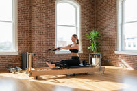 A woman in black athletic wear seated on a foldable oak pilates reformer with metal frames and adjustable cables uk delivery