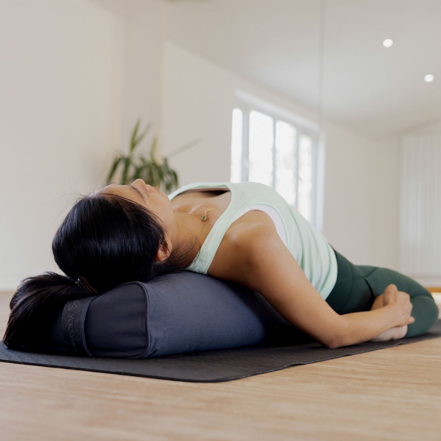 A woman in a yoga pose lying on a dark gray bolster pillow on a black mat in a bright, modern studio with light wood floors.